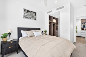 Quarto moderno com cama grande, mesas de cabeceira, guarda-roupa e área de cozinha visível por uma porta aberta.