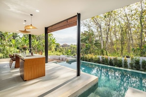 Un área de piscina exterior moderna con bar y asientos, rodeada de exuberante vegetación y árboles.