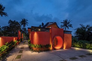 Un edificio moderno con una ventana circular distintiva y un techo inclinado, rodeado de abundante vegetación y palmeras.