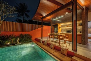 A modern outdoor pool area with a dining setup and a kitchen visible through glass doors.
