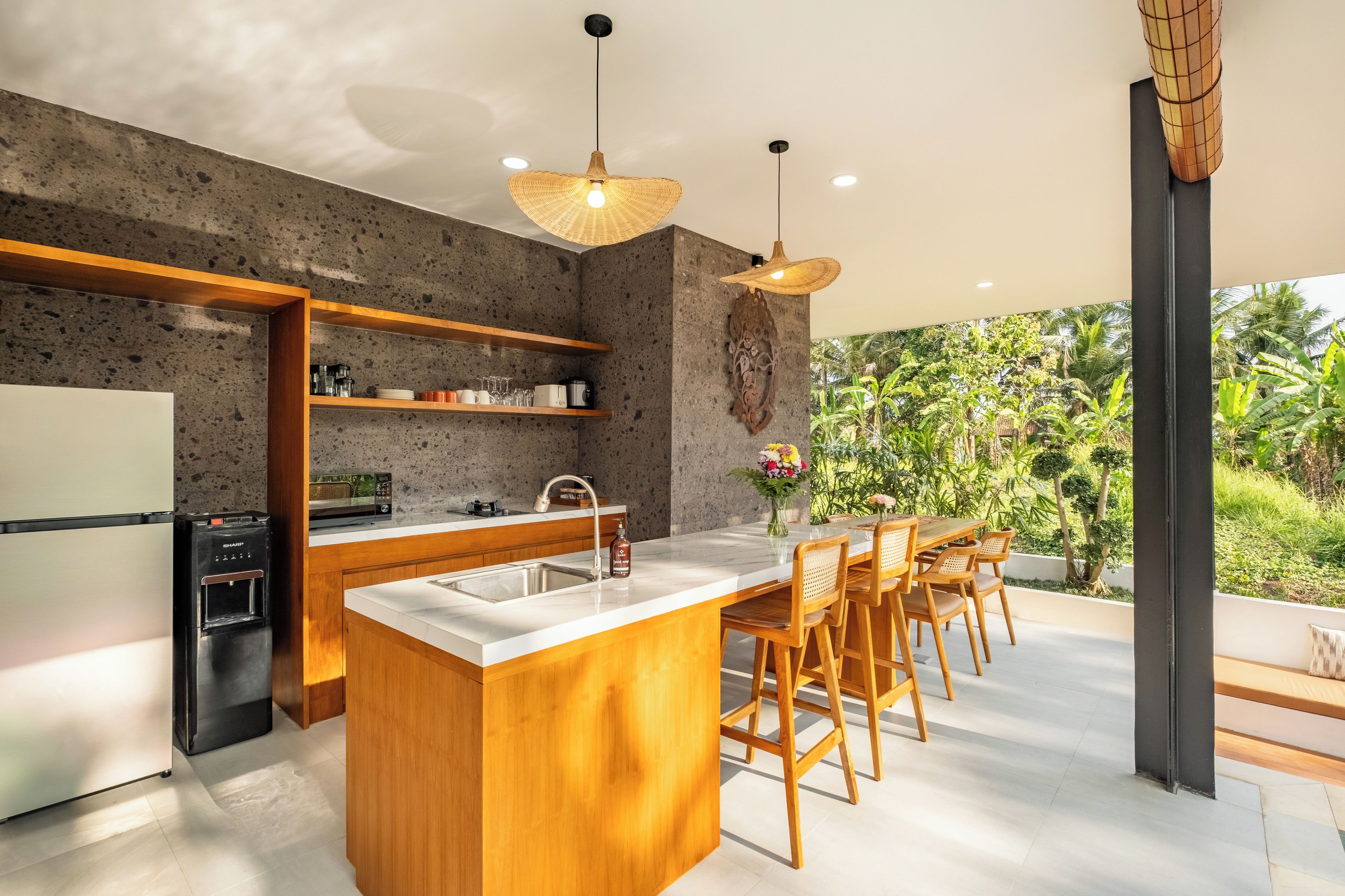 A modern kitchen with a central island, wooden bar stools, and a view of lush greenery.