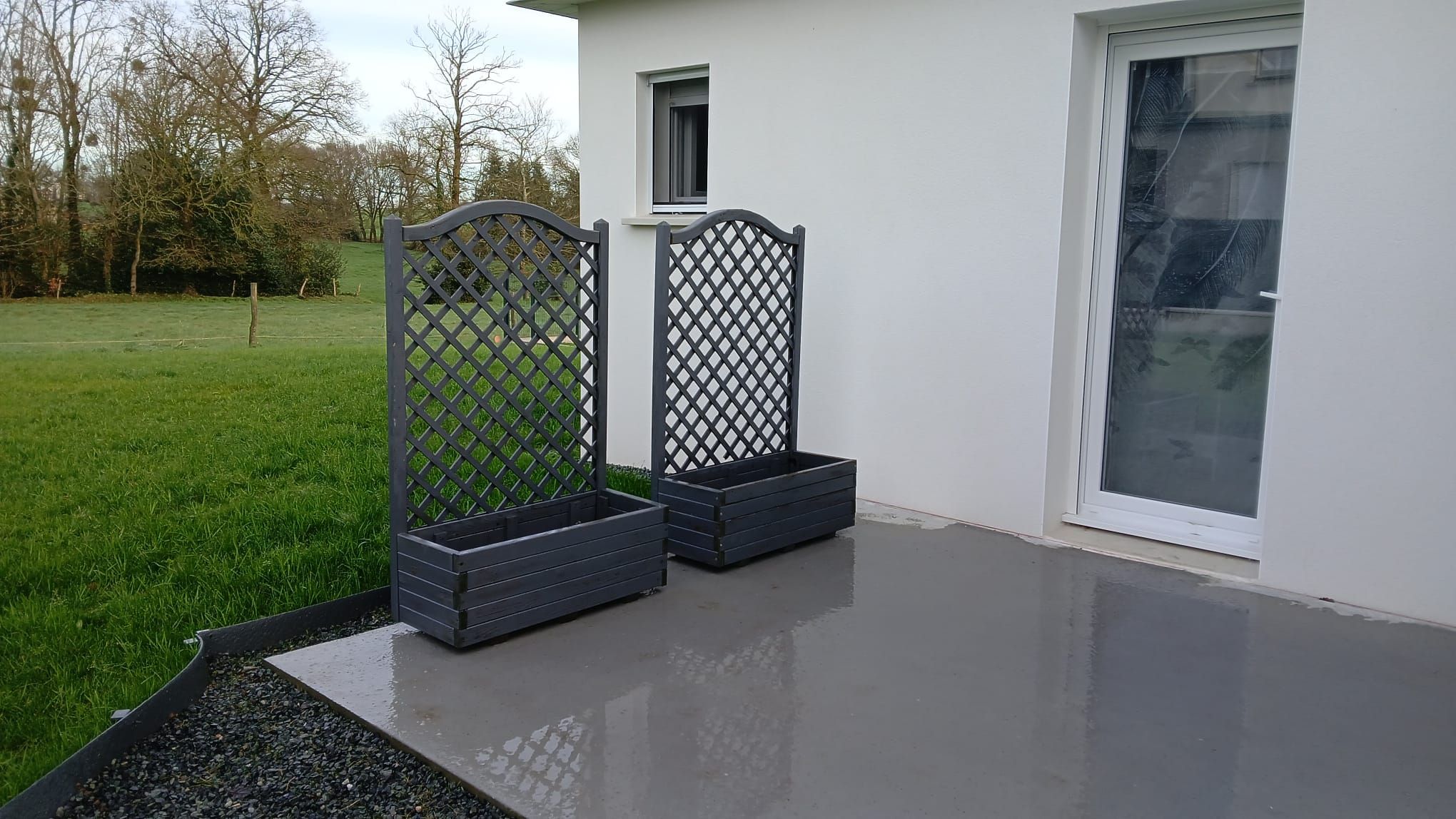 Een modern buitengebied met een betonnen vloer, twee donkerhouten plantenbakken en een wit gebouw met een raam en een deur.