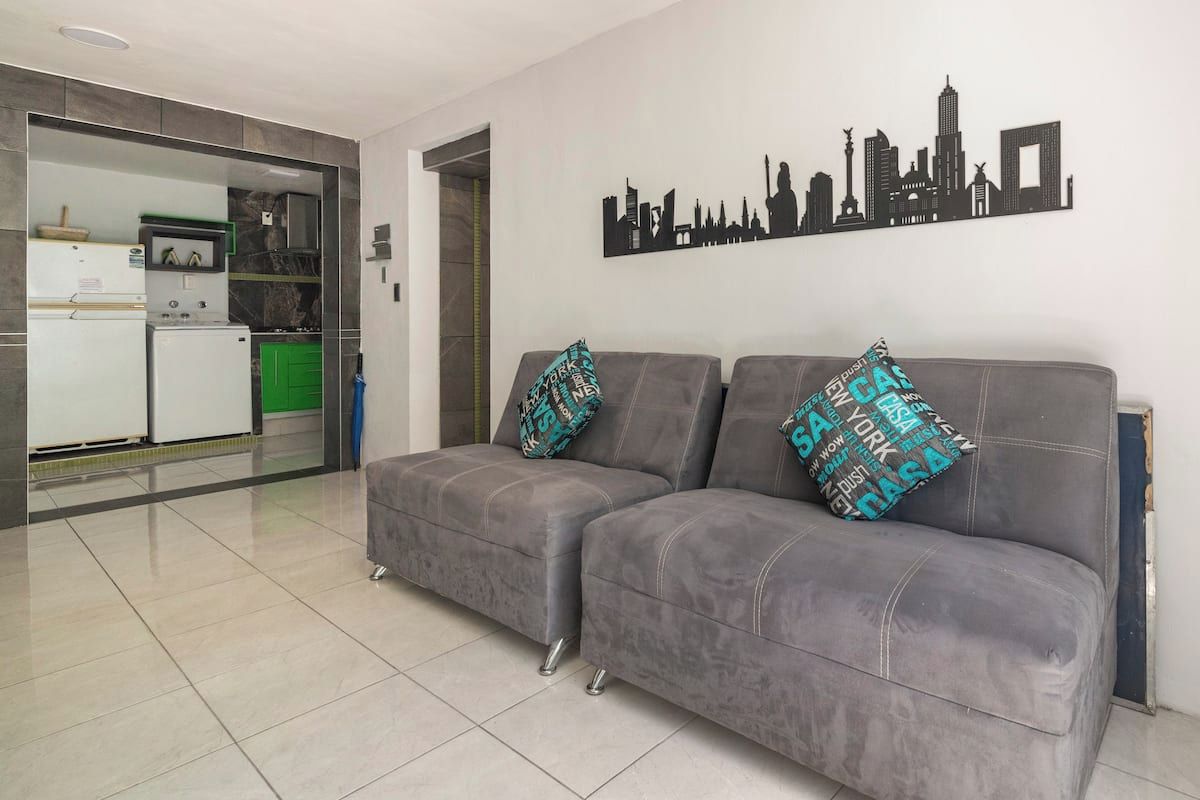Un soggiorno con un divano grigio, due cuscini abbinati, un quadro raffigurante uno skyline cittadino e una cucina visibile tramite un'apertura.