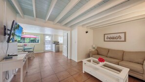 A living room with a beige sofa, a white coffee table, and a TV on a stand. There is a dining area with a table and chairs, and a kitchen visible in the background. - Pool, Spacious House, Close to Siesta and Downtown (Sarasota)