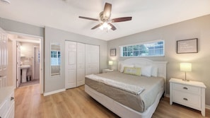 Une chambre à coucher comprenant un lit, des tables de chevet, un ventilateur de plafond, une fenêtre et une porte donnant sur une salle de bain.