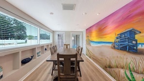 Un espace repas avec une longue table et des chaises, une fresque représentant une scène de plage et une vue dégagée grâce à de grandes fenêtres.