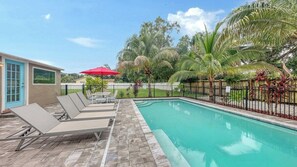 Une piscine entourée de chaises longues et d’un parasol rouge, le tout bordé de palmiers et délimité par une clôture.