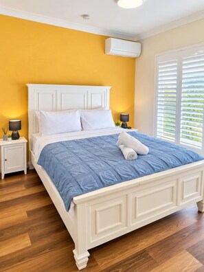 A bedroom with a white bed, blue bedding, a white nightstand with a lamp, and a window with blinds.