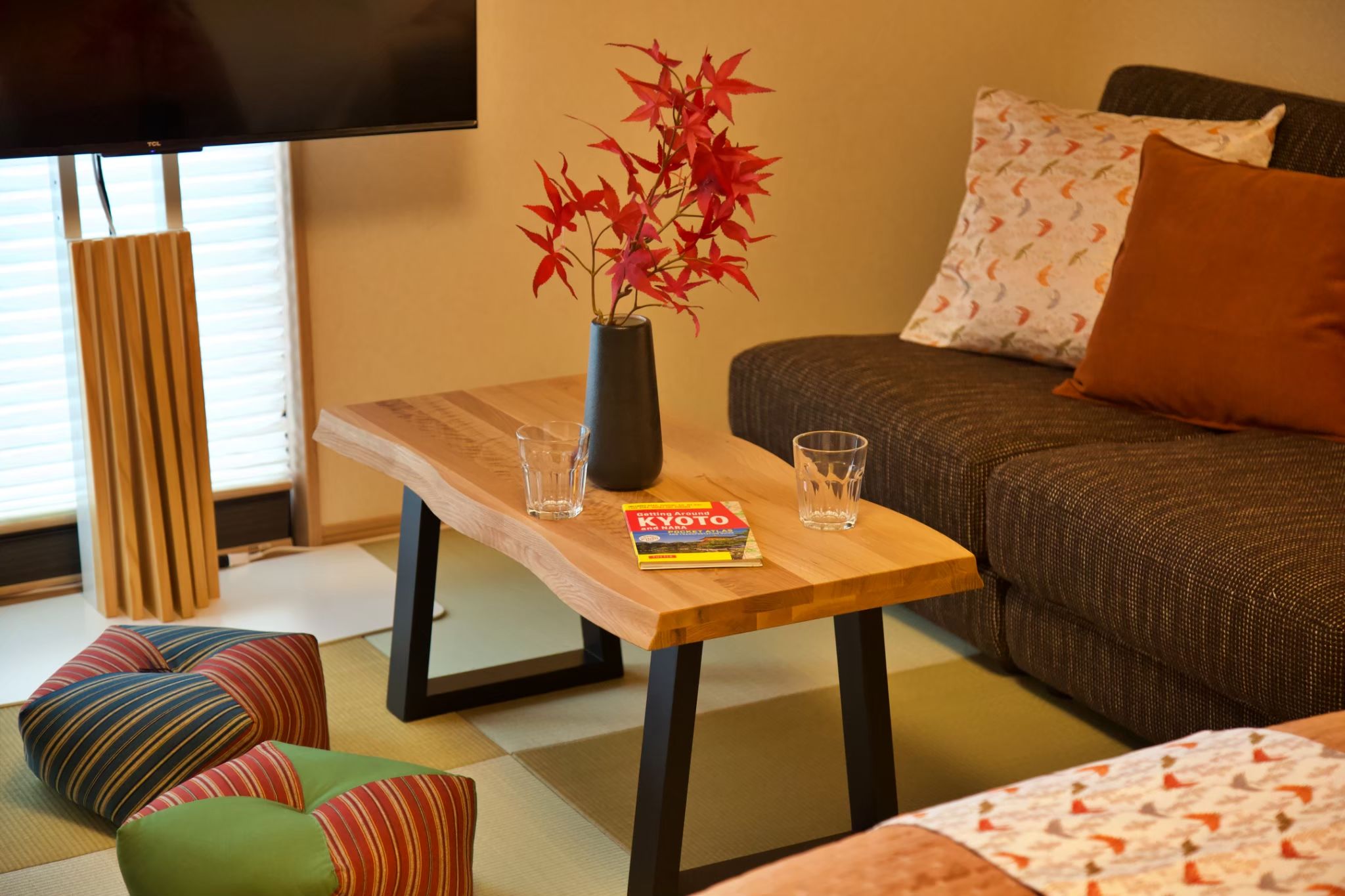A living room with a wooden table, a vase with red leaves, a book, and a sofa with patterned cushions.
