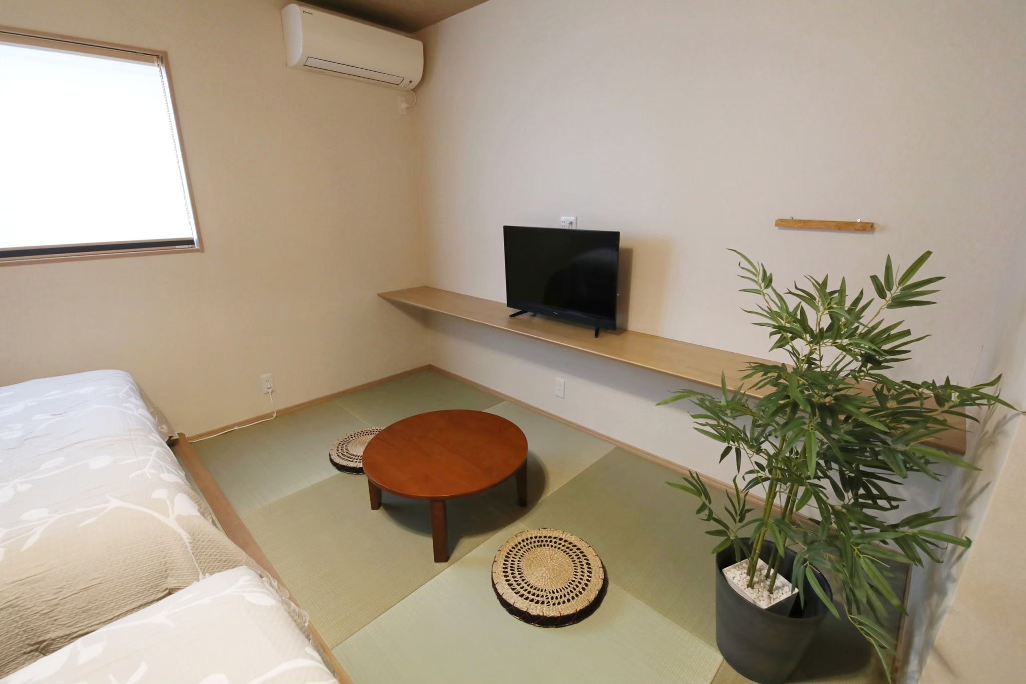 A room with tatami flooring, a bed, a television on a shelf, a small round table, and a potted plant.
