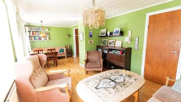 Een gezellige woonkamer met een bank, fauteuil en een salontafel. Er staat een boekenplank vol boeken en er is een deur aan de rechterkant.