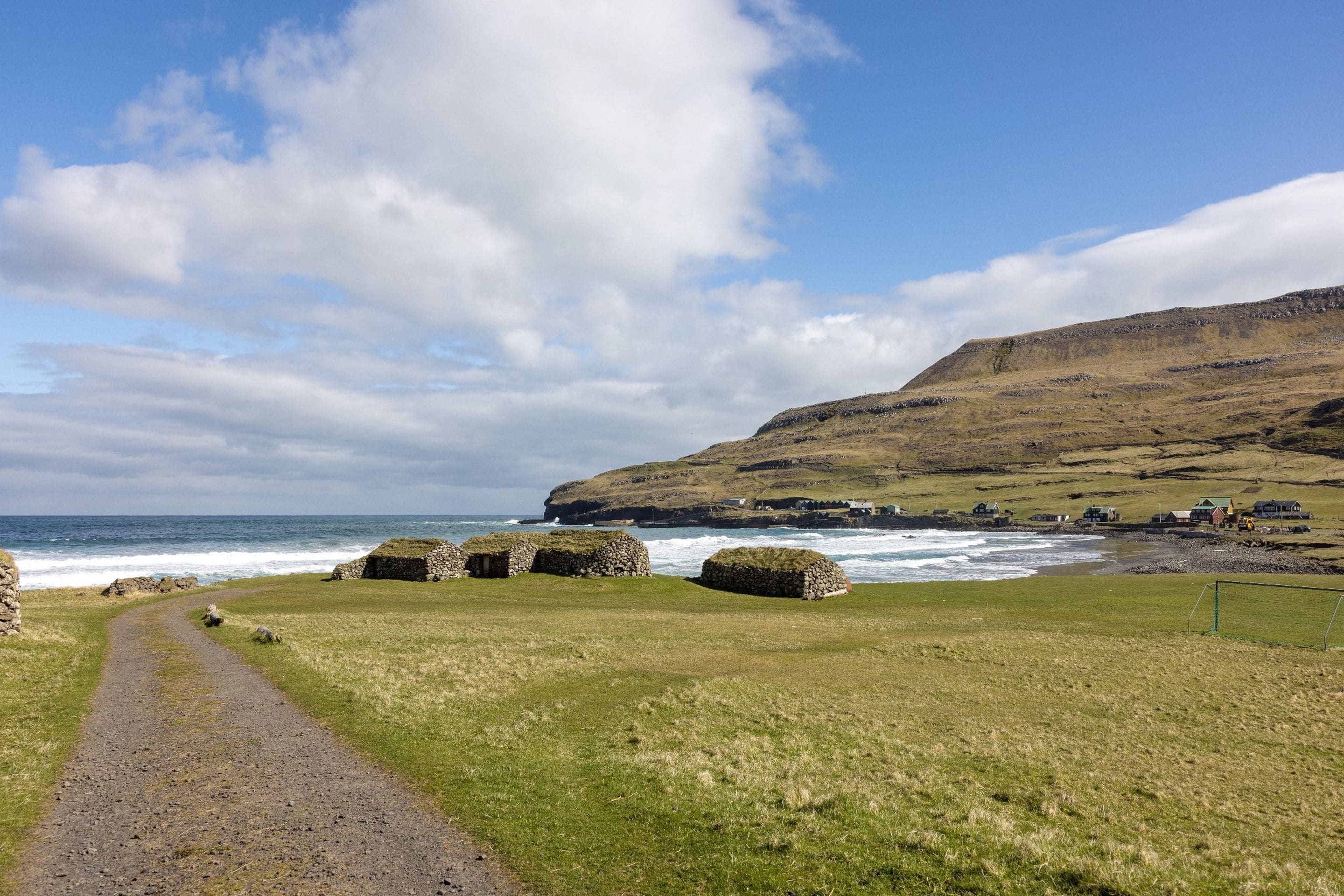 Ein Küstenpfad mit Steinbauten, einer grünen Fläche und Blick auf Meer und Hügel.