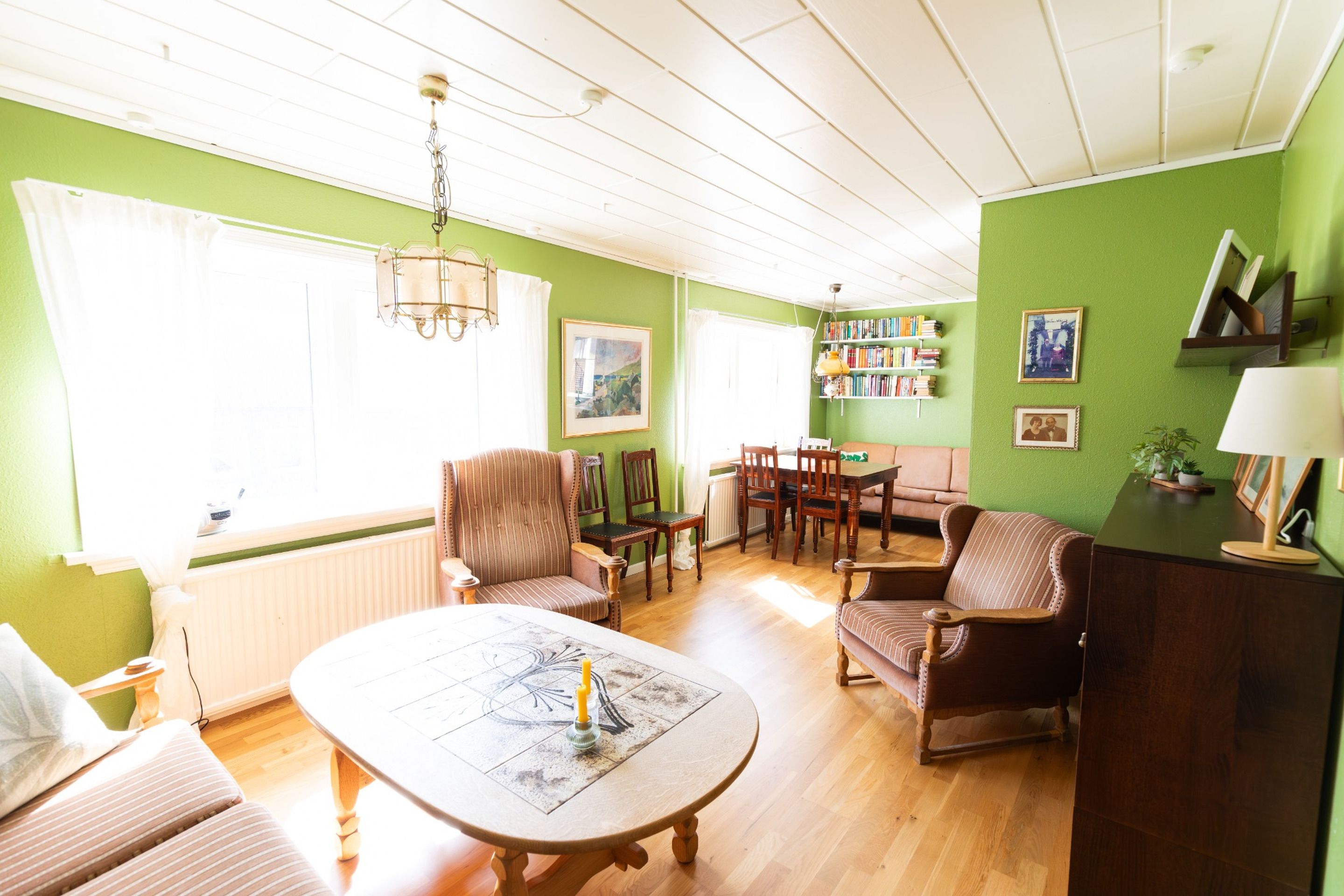 A well-lit living room with a dining area, green walls, and a bookshelf.