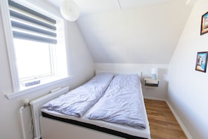 Una habitación pequeña con cama individual, techo inclinado, ventana con persianas, mesita de noche con lámpara y radiador.