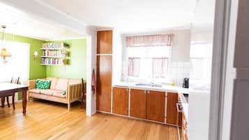 A bright living room with a wooden floor, a sofa, a dining table, and a kitchen area with wooden cabinets and a window with checkered curtains.