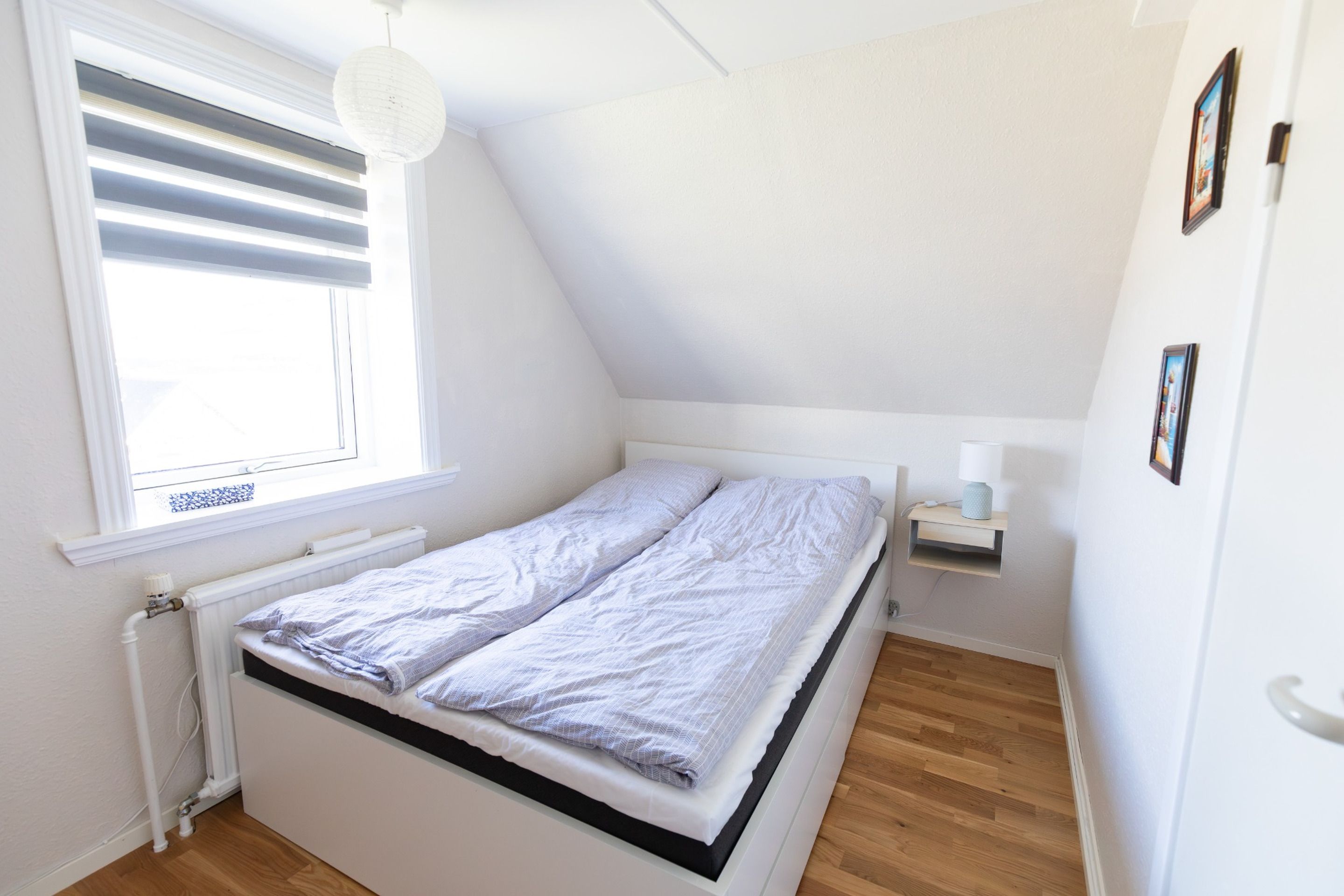 A small, well-lit bedroom with a single bed, a radiator, a window with blinds, and a wall-mounted shelf.
