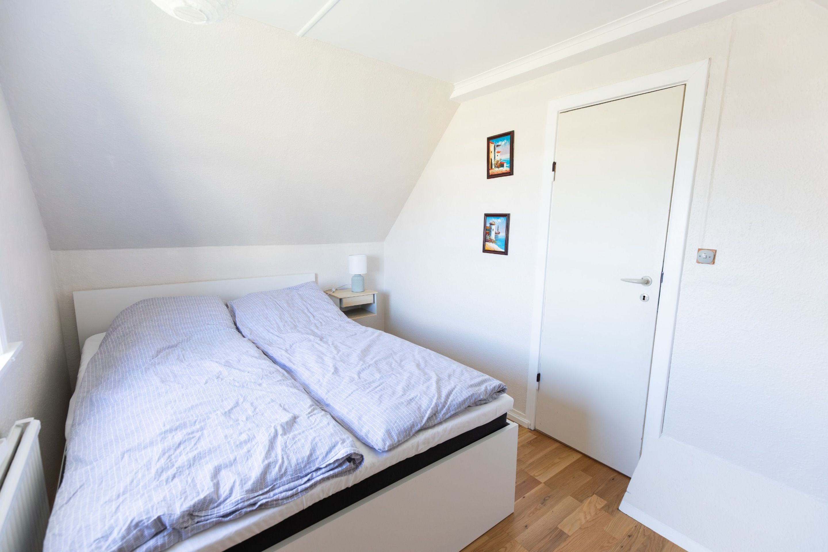 A small, single-bed room with a sloped ceiling, a bedside table with a lamp, and two framed pictures on the wall.