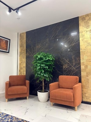 A room with two orange armchairs, a potted plant, a marble wall, and a framed picture on the wall. - Avenue Hotel (Almaty)