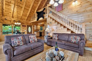 A log cabin interior with two leather sofas, a dining area, a kitchen, and a staircase with a black bear decoration. - 5-Acre Smoky Cabin • Hot Tub + Mountain Views • Near Pigeon Forge | Eagle’s View (Sevierville)