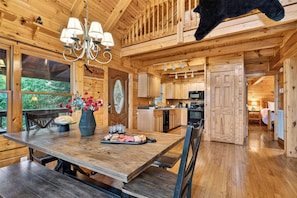 A wooden cabin interior with a dining area, kitchen, and a view of the outdoors. - 5-Acre Smoky Cabin • Hot Tub + Mountain Views • Near Pigeon Forge | Eagle’s View (Sevierville)