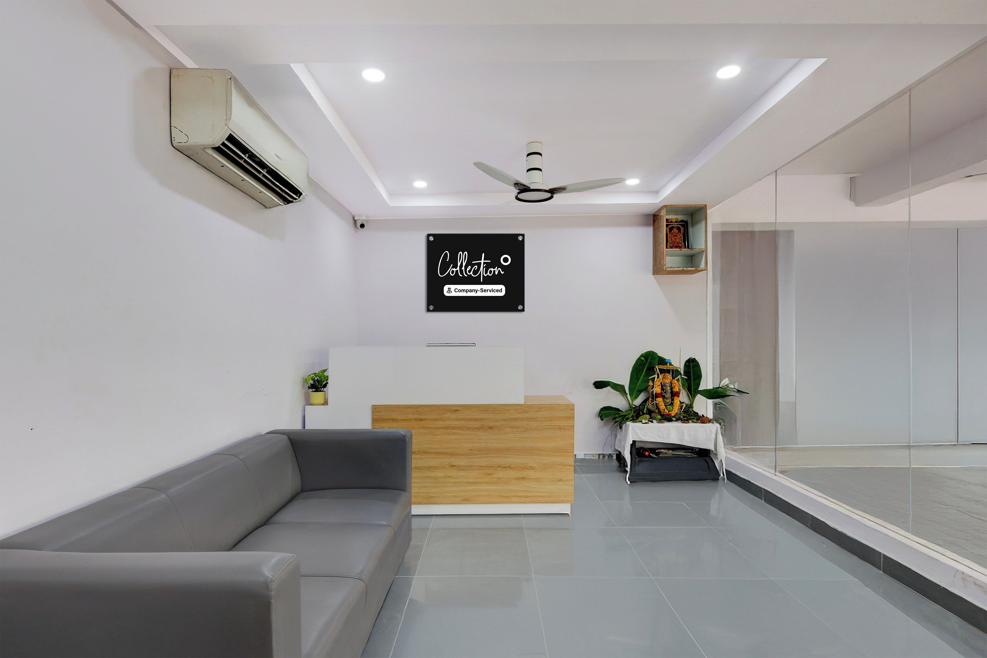 A reception area with a grey sofa, a wooden reception desk, a ceiling fan, and a sign indicating the space is company-serviced.