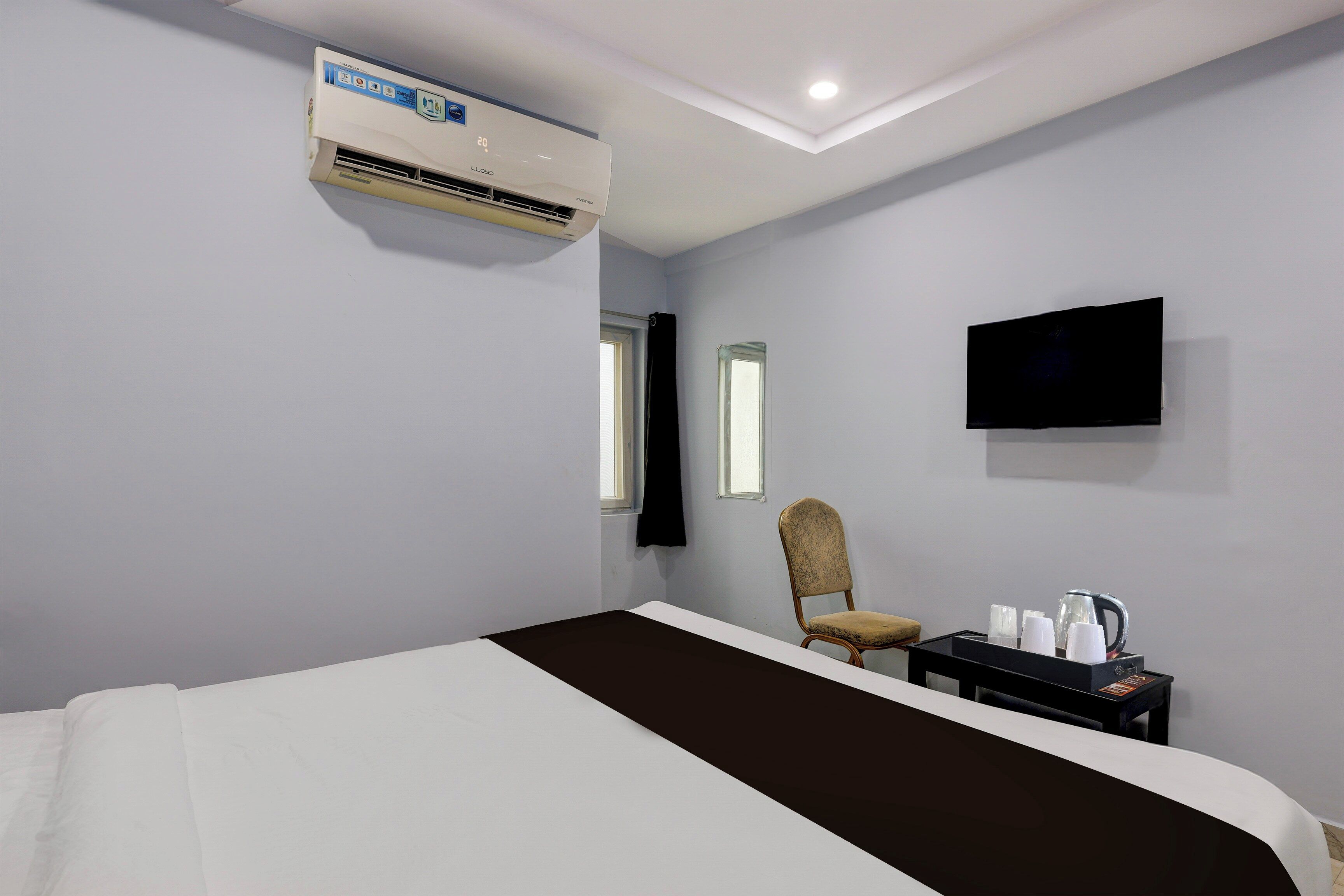 A hotel room with a bed, a chair, a table with a kettle, a flat-screen TV, and an air conditioning unit.