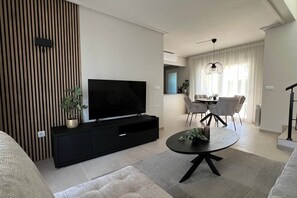 A modern living room with a flat-screen TV on a black entertainment unit, a round coffee table with a plant, and a dining area with a chandelier. - Villa Olivia - Mar Menor Golf Resort (Torre-Pacheco)