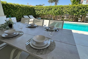 Une table à manger dressée avec des assiettes, des bols et des verres à vin, donnant sur une piscine.