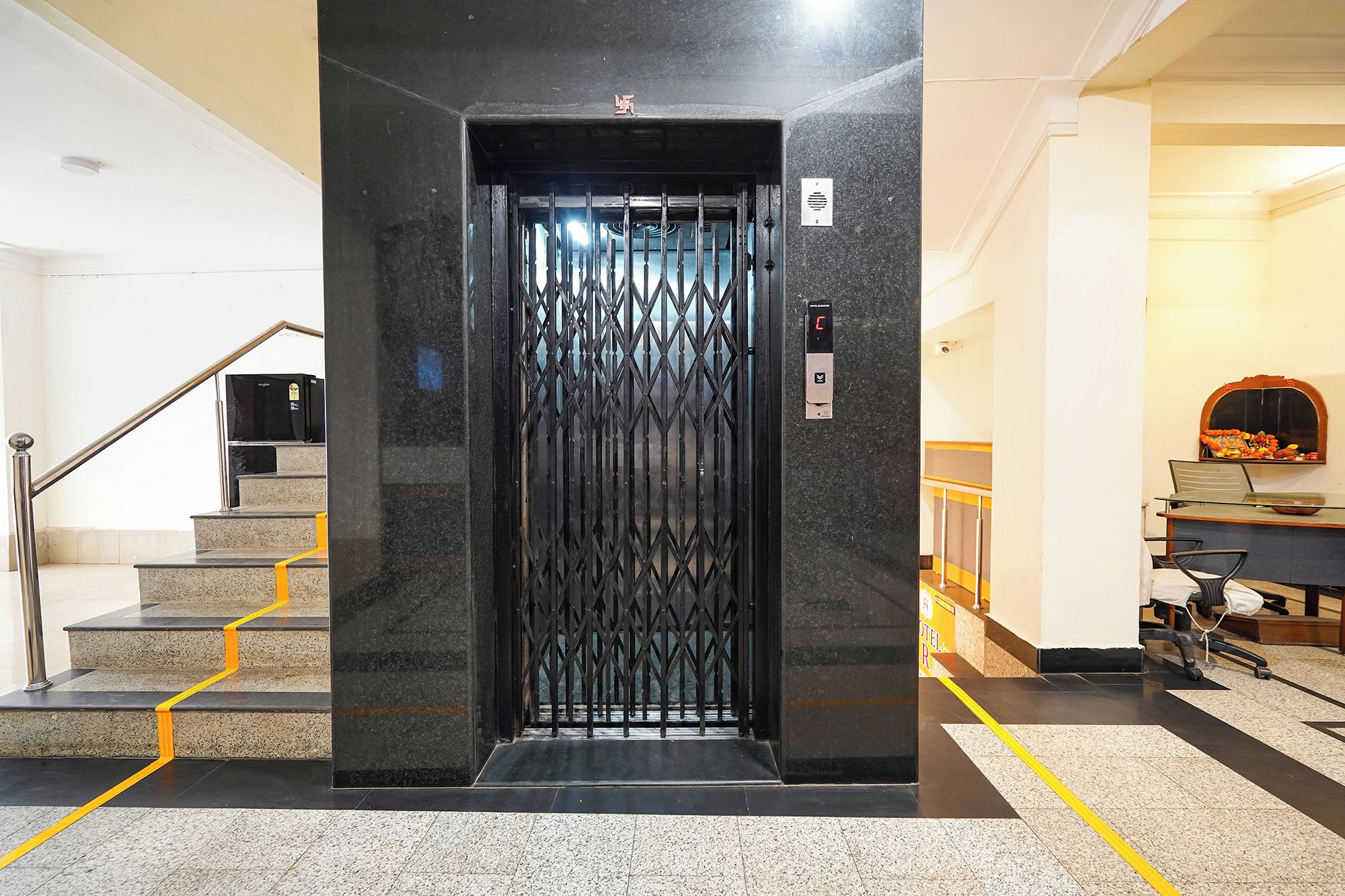 Un ascensor con celosía metálica, una escalera con barandilla y una zona de recepción con mostrador y sillas.