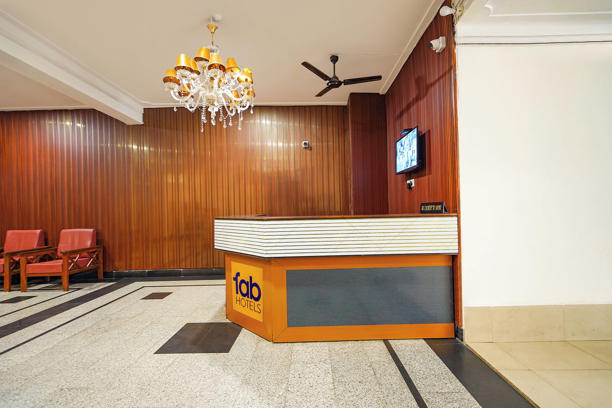 Zona de recepción de un hotel con candelabro, mostrador de recepción, televisor y área de descanso con sillones.