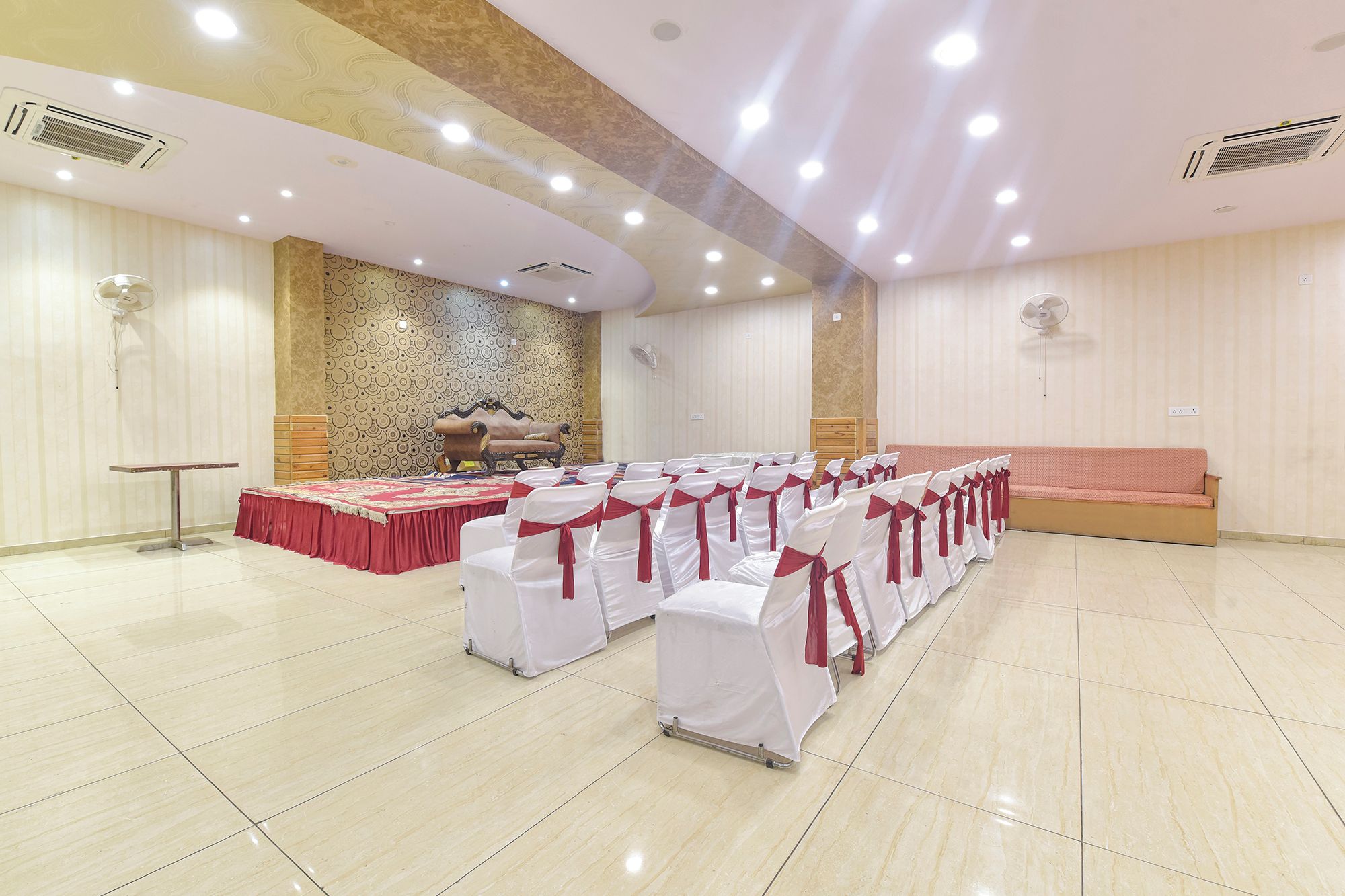 Un salón de banquetes con filas de sillas blancas con lazos rojos, un escenario con mantel rojo y un sofá.