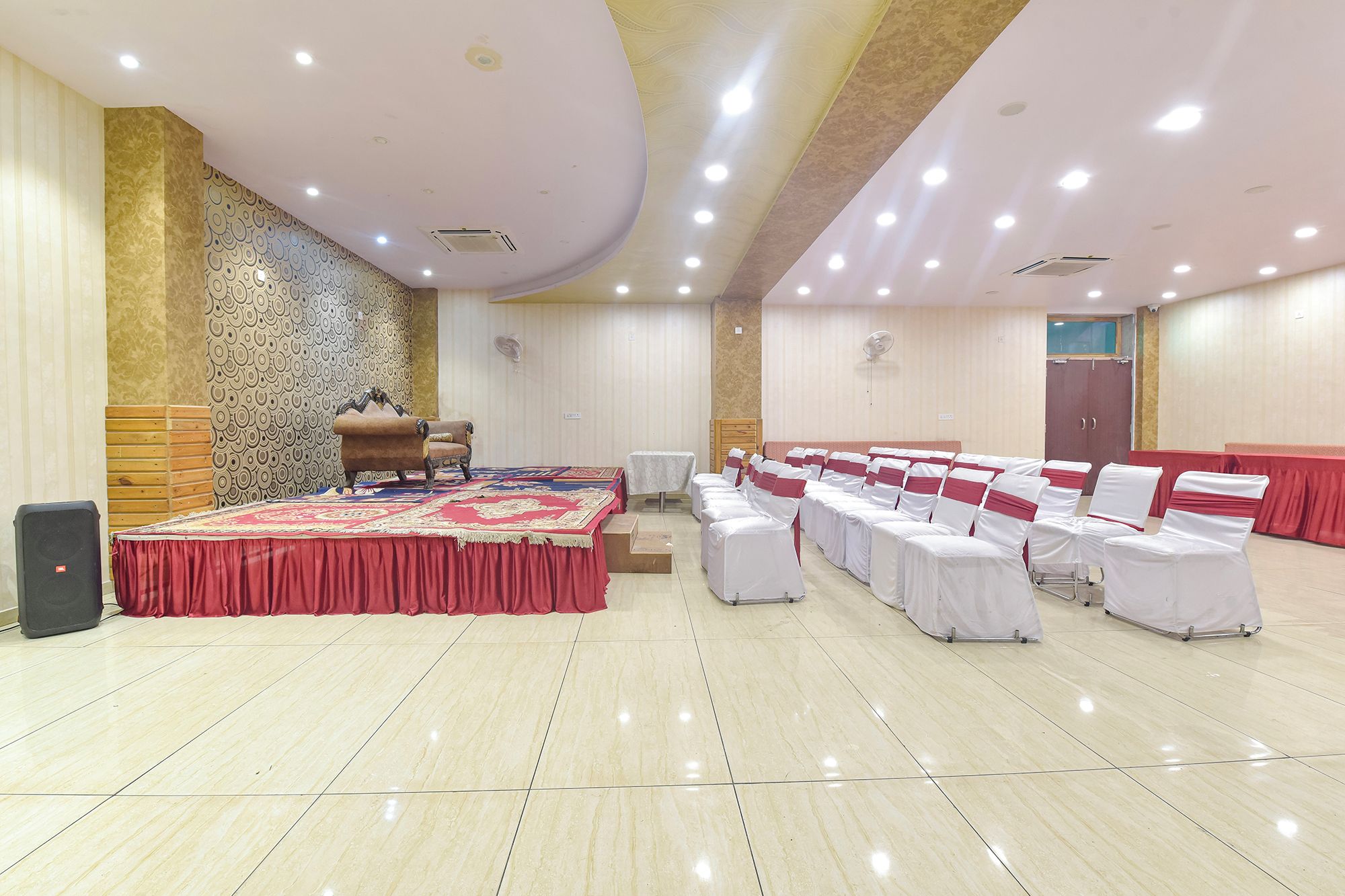 Un salón de conferencias con un escenario, filas de sillas blancas con cintas rojas, una alfombra roja grande y un muro con diseño.