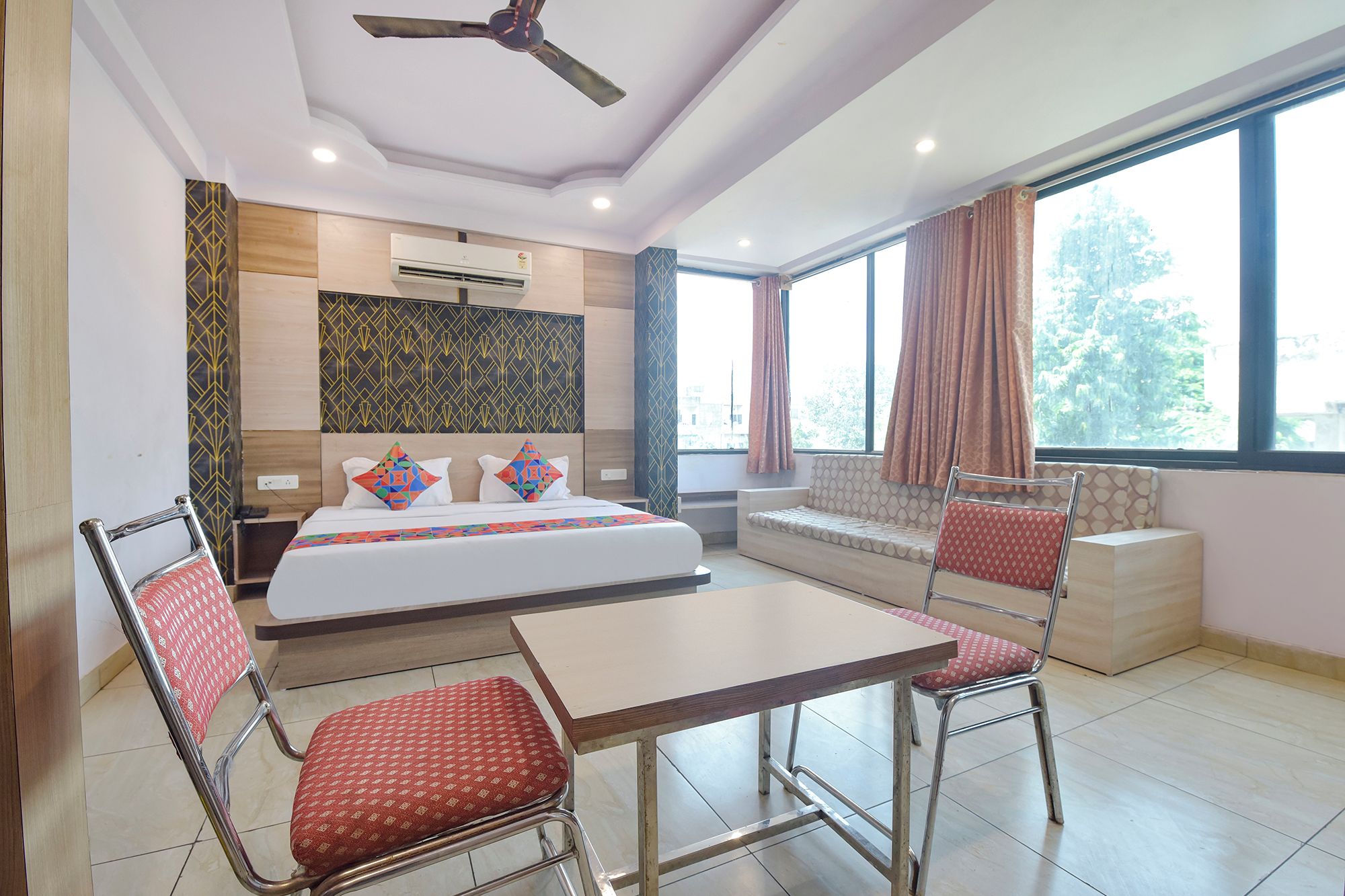 A hotel room with a bed, a desk, two chairs, a ceiling fan, and large windows.