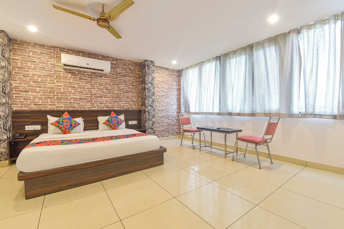 A hotel room with a brick wall, a bed with colorful pillows, a ceiling fan, an air conditioning unit, a window with sheer curtains, and a small table with chairs.