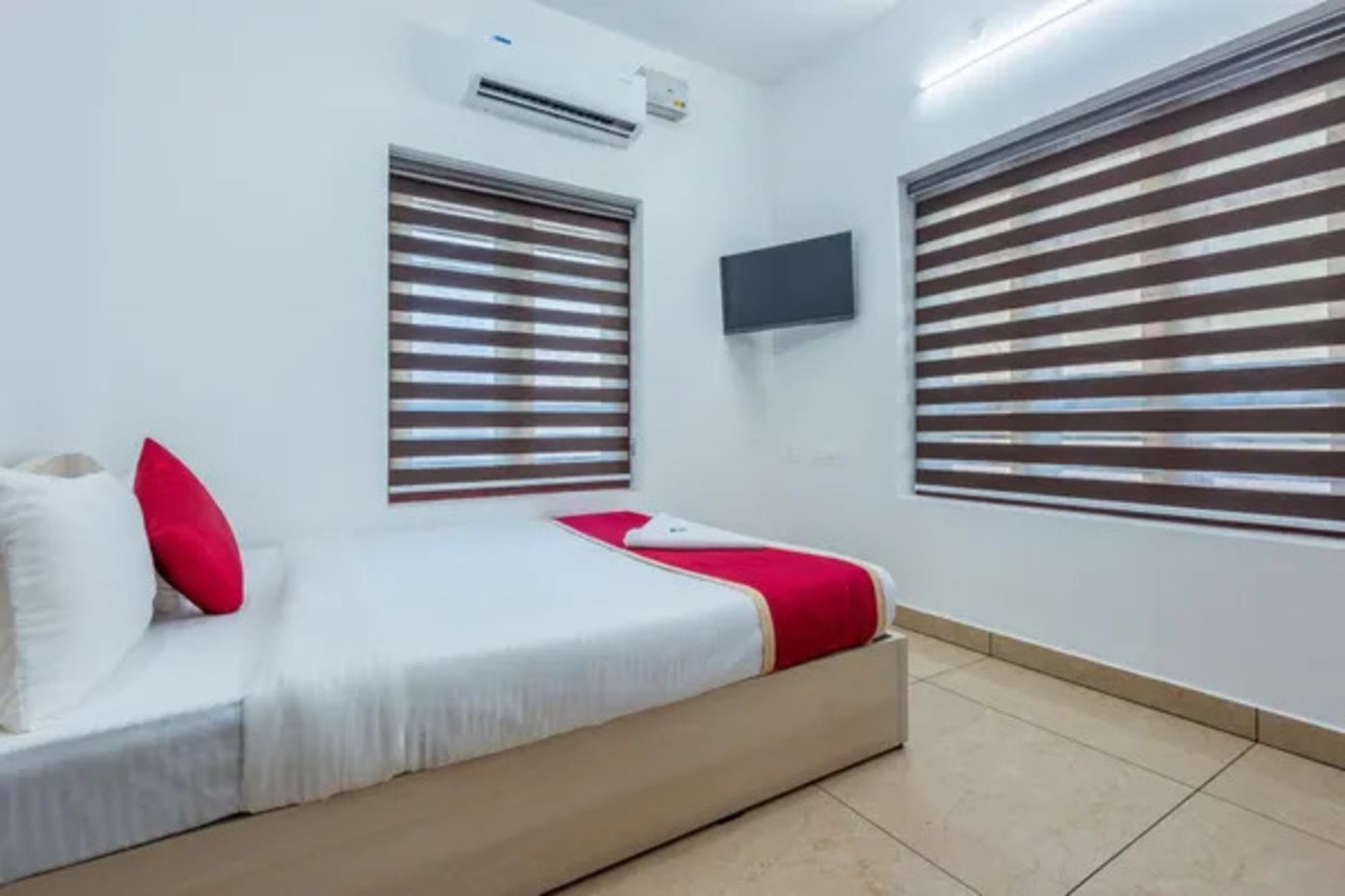 A hotel room with a bed, two windows with blinds, a flat-screen TV, and an air conditioning unit.