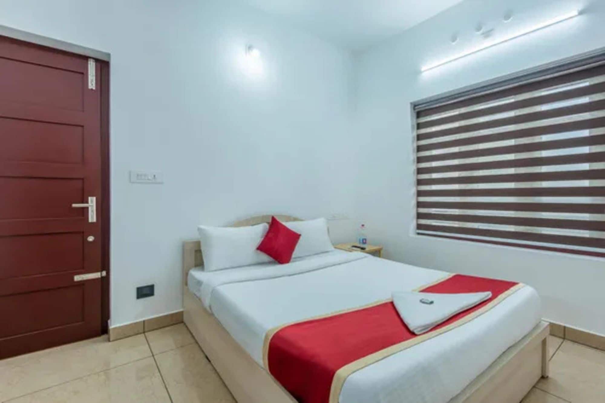 A hotel room with a bed, a nightstand, a red door, and a window with blinds.