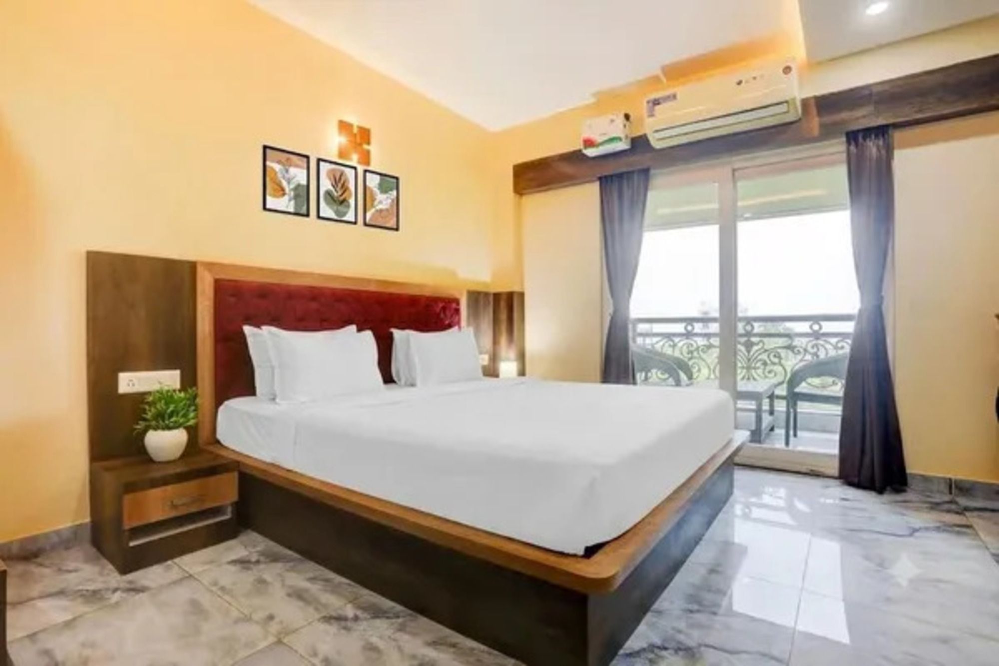 A hotel room with a large bed, a bedside table with a plant, a wall-mounted air conditioner, and a balcony with a view.