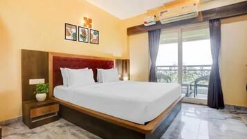 A hotel room with a large bed, a bedside table with a plant, a wall-mounted air conditioner, and a balcony with a view.