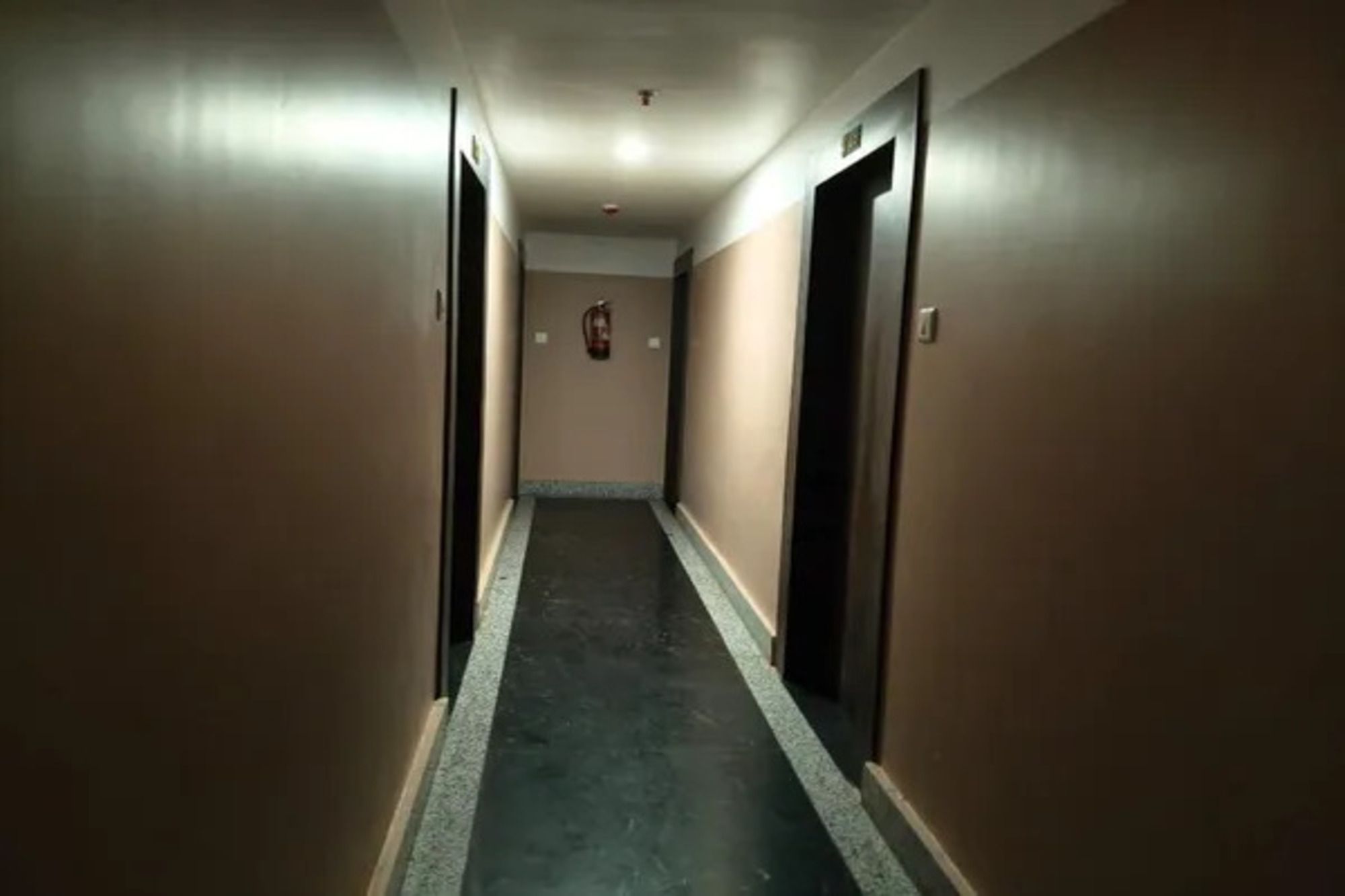 A corridor with doors on both sides, a fire alarm on the wall, and a ceiling with recessed lighting.