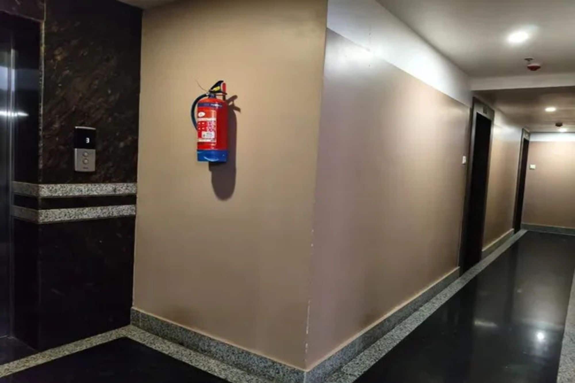 Un pasillo con un extintor montado en la pared, una puerta a la izquierda y una lámpara en el techo.
