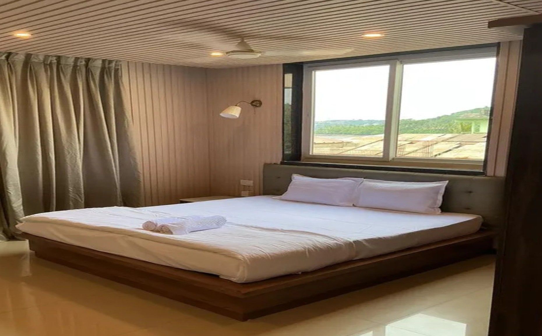 Ein ordentlich bezogenes Bett in einem Zimmer mit großem Fenster, Vorhängen und einer Decke mit Rastermuster.