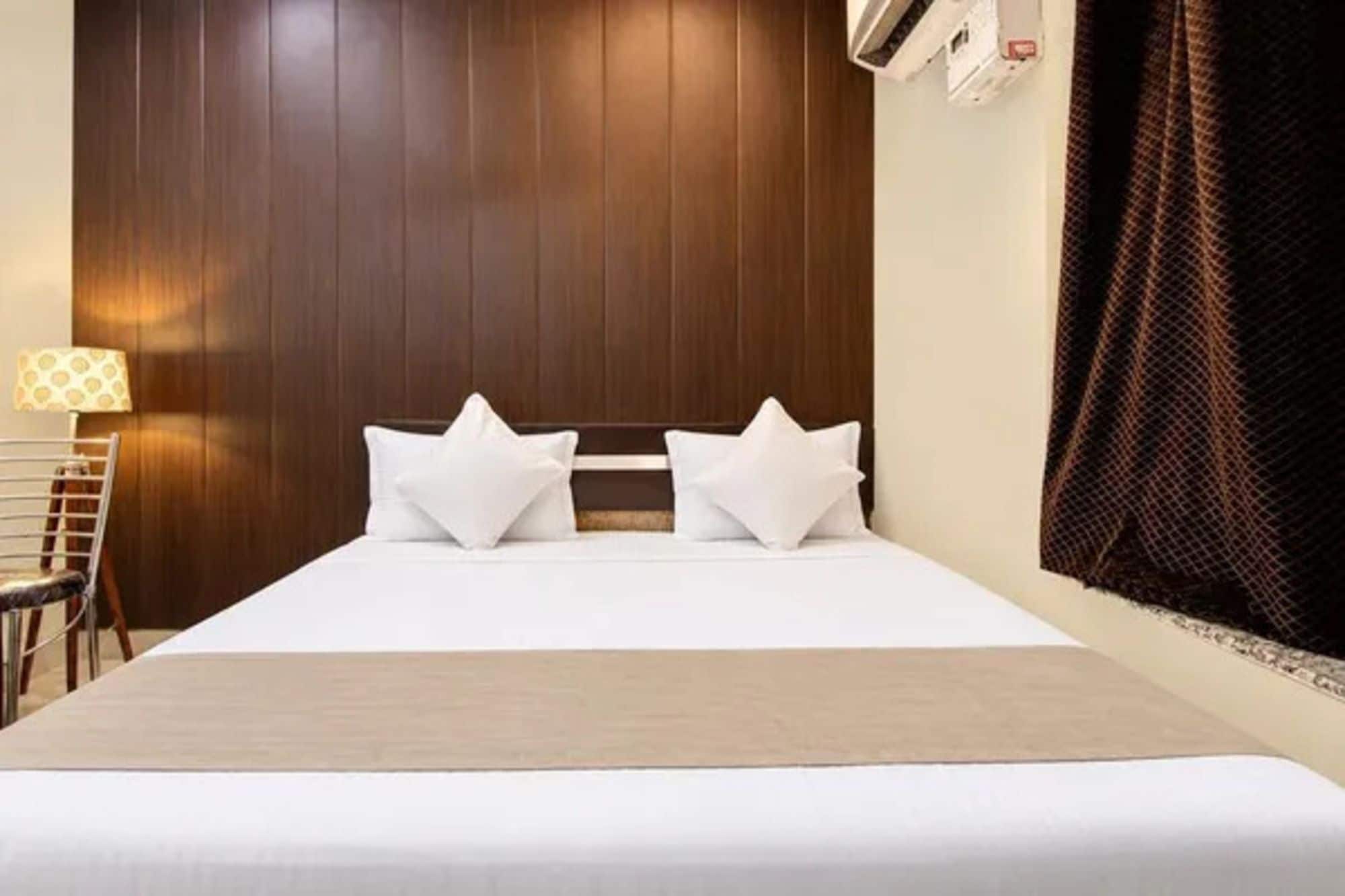 Habitación de hotel con cabecera de madera, cama con ropa blanca, dos almohadas blancas, una mesita de noche y una lámpara.