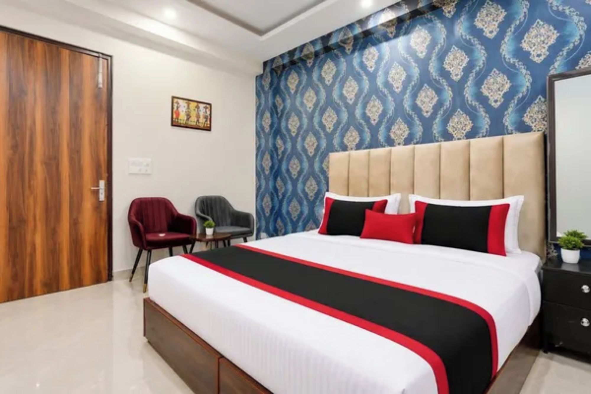A hotel room with a large bed, a headboard, a red armchair, a grey armchair, a small table, and a wall with a patterned wallpaper.