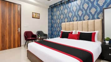 A hotel room with a large bed, a headboard, a red armchair, a grey armchair, a small table, and a wall with a patterned wallpaper.