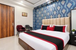 Habitación de hotel con una cama grande, cabecera, un sillón rojo, un sillón gris, una mesita y una pared con papel tapiz estampado.