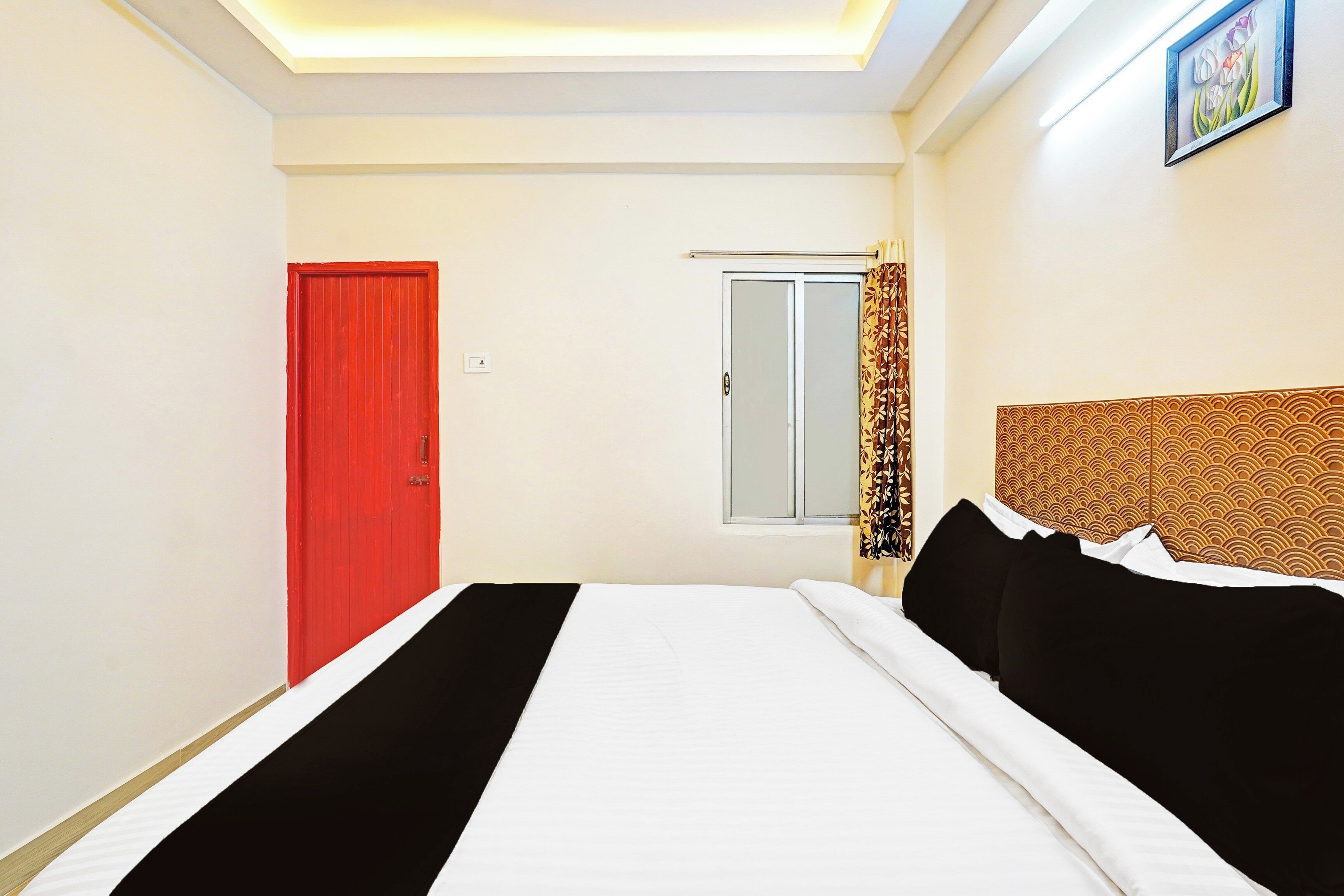 Ein Hotelzimmer mit einem Bett, einer roten Tür, einem Fenster mit Vorhang und einem an der Wand hängenden Bild.