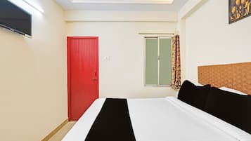 A hotel room with a bed, a red door, a window with curtains, and a wall-mounted television.