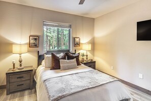 Ein Schlafzimmer mit einem Bett, zwei Nachttischen, einem Fenster mit Blick auf Bäume und einem an der Wand montierten Fernseher.