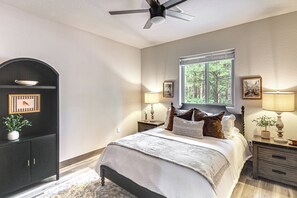 A bedroom with a bed, nightstands, a ceiling fan, a window with a view of trees, and a dark arched cabinet with a framed picture. - Golfer’s Oasis! Ponderosa Pine Retreat in Show Low (Show Low)