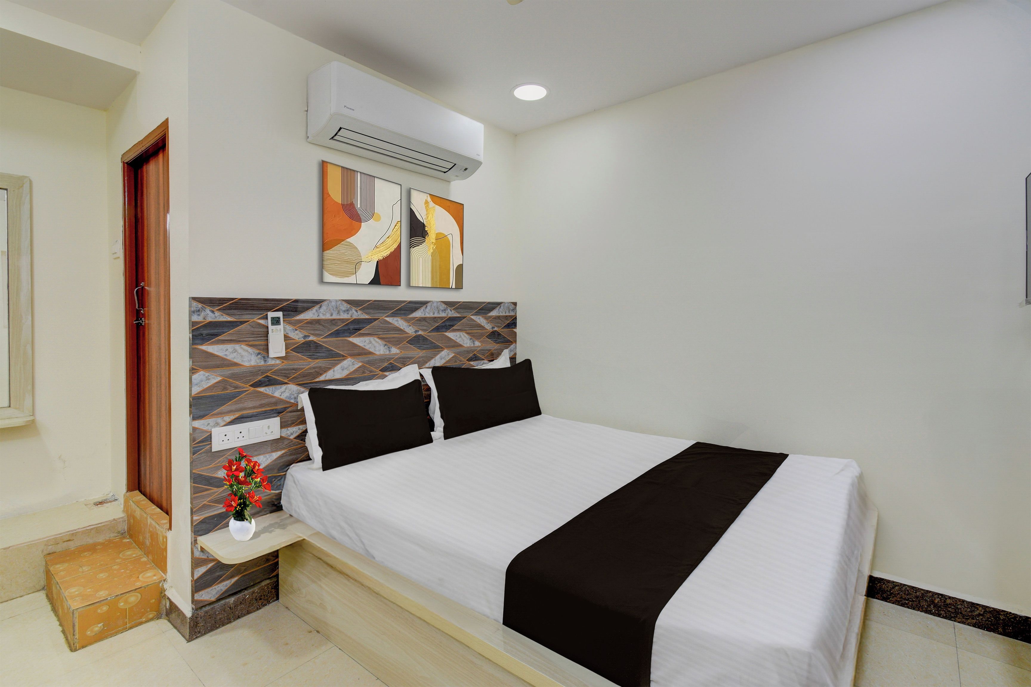 Habitación de hotel con una cama, una mesita de noche, un aire acondicionado de pared y dos cuadros abstractos.